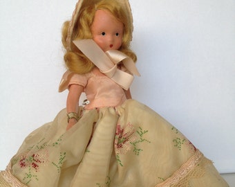 Puede incluir: Muñeca vintage con cabello rubio, vestida con un vestido rosa y un gorro a juego. El vestido de la muñeca presenta una falda color crema con bordados florales y un ribete de encaje. La muñeca es un objeto de colección.