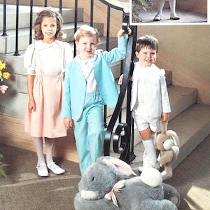 Könnte beinhalten: Drei Kinder in Osteroutfits posieren auf einer Treppe. Ein Mädchen trägt ein rosa Kleid mit weißen Schuhen. Ein Junge trägt einen hellblauen Anzug, und ein anderer Junge trägt ein weißes Outfit und hält einen Stoffhasen. Ein großer grauer Stoffhase liegt auf der Treppe.