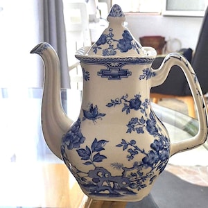 Może przedstawiać: Biały ceramiczny imbryk z niebieskim kwiatowym wzorem. Imbryk ma zakrzywioną wylewkę, uchwyt i pokrywkę z guzikiem. Niebieski kwiatowy wzór zawiera kwiaty, liście i inne elementy dekoracyjne.