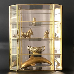 May include: A hexagonal glass display cabinet with a gold-toned metal frame. The cabinet features multiple glass shelves containing various brass figurines, including a dolphin, a small bowl with handles, a horse, and other small decorative items.
