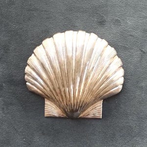 May include: A decorative, metallic seashell-shaped object. The shell has a fan-like design with textured ridges and a square base. The object is a light gold colour and sits against a dark grey background.
