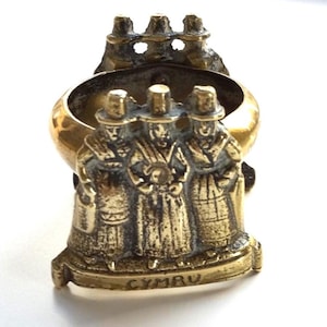 May include: A vintage brass ring featuring three figures in traditional Welsh attire. The ring has a gold-colored band and a detailed base with the word "CYMRU" inscribed. The figures are wearing hats and long coats.