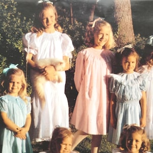 Könnte beinhalten: Eine Gruppe junger Mädchen in Vintage-Kleidern posiert im Freien. Ein Mädchen hält eine flauschige, weiße Katze. Die Kleider sind in Weiß-, Rosa- und Blautönen gehalten, mit passenden Schleifen im Haar. Der Hintergrund zeigt Grün und einen Baumstamm.