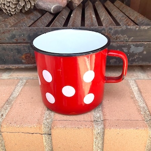 Könnte beinhalten: Eine rote Emaille-Tasse mit weißen Tupfen. Die Tasse hat einen schwarzen Rand und Henkel sowie eine weiße Innenseite. Die Tasse steht auf einer Ziegeloberfläche.