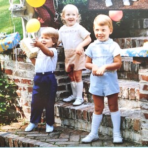 Könnte beinhalten: Drei Kinder in Vintage-Kleidung stehen auf Backsteinstufen. Eines hält ein Geschenk und einen gelben Ballon. Die Kinder lächeln. Die Szene deutet auf eine Geburtstagsfeier mit festlicher Atmosphäre hin.