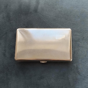 Könnte beinhalten: Eine rechteckige, silberfarbene Zigarettenetui mit einem strukturierten, kreuzschraffierten Muster auf dem Deckel. Die Hülle hat einen kleinen, rechteckigen Verschluss am unteren Rand. Die Hülle ist geschlossen und liegt auf einer dunkelgrauen Oberfläche.