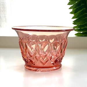 Könnte beinhalten: Eine rosa Glasschale mit einem dekorativen Muster. Die Schale hat einen breiten, leicht ausgestellten Rand und ein detailliertes, strukturiertes Design auf der Außenseite. Die Schale ist wahrscheinlich Vintage und eignet sich für die Präsentation oder zum Servieren.