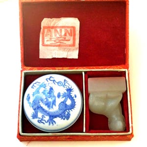 Peut inclure: Boîte rouge ouverte contenant un sceau chinois. L'ensemble comprend un tampon encreur rond blanc avec un motif de dragon bleu, un sceau en pierre transparente et un tampon encreur rouge avec les lettres "ANN".