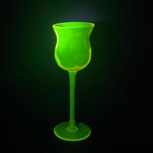 Op de afbeelding: Een gloeiende neon-groene glazen kelk met een tulpvormige kom, een slanke steel en een ronde voet. Het glas straalt een helder, lichtgevend schijnsel uit tegen een donkere achtergrond, wat de gladde, gebogen vorm en doorschijnende textuur benadrukt.