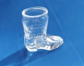 May include: A clear glass boot-shaped container with the word "BEA" visible on the side. The boot has a rounded top and a detailed design, sitting on a blue surface. The container is likely a decorative item or a small vase.