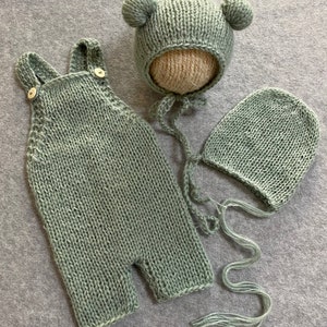 May include: A set of three knitted baby clothes in a sage green color. The set includes a pair of overalls, a bonnet with bear ears, and a beanie.
