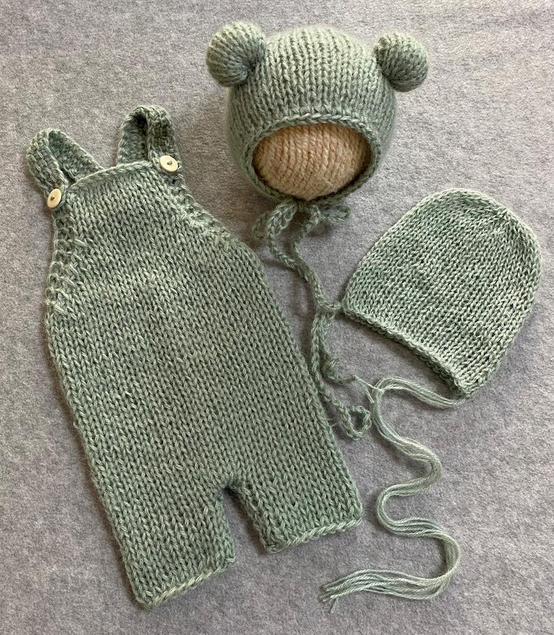 May include: A set of three knitted baby clothes in a sage green color. The set includes a pair of overalls, a bonnet with bear ears, and a beanie.