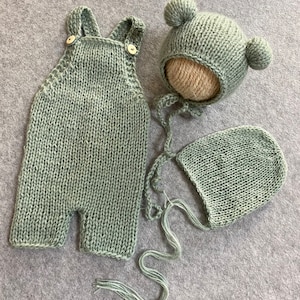 May include: A sage green knitted baby outfit set with a hat and a bonnet. The hat has two pom-poms on top.