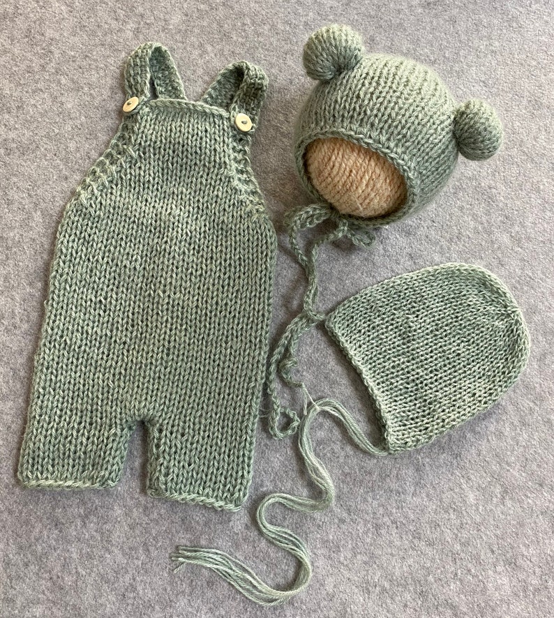 May include: A sage green knitted baby outfit set with a hat and a bonnet. The hat has two pom-poms on top.