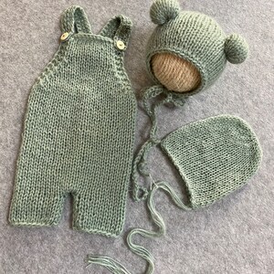 May include: A sage green knitted baby outfit set with a hat and a bonnet. The hat has two pom-poms on top.