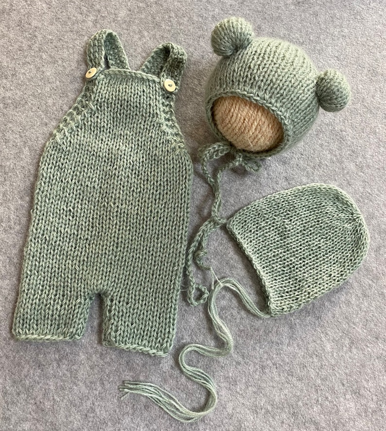 May include: A sage green knitted baby outfit set with a hat and a bonnet. The hat has two pom-poms on top.