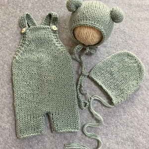 May include: A sage green knitted baby outfit set with a hat and a bonnet. The hat has two small pom-poms on top.