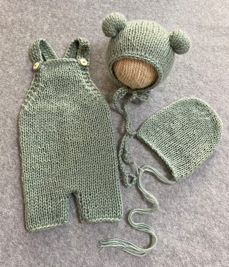 May include: A sage green knitted baby outfit set with a hat and a bonnet. The hat has two small pom-poms on top.