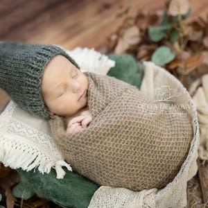 Può includere: Un neonato dorme pacificamente avvolto in una coperta di maglia marrone, indossando un berretto a punta di maglia verde. Il bambino è sdraiato su uno sfondo di tessuto verde e bianco.