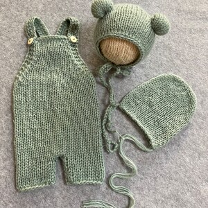 May include: A sage green knitted baby outfit set with a hat, a bonnet, and overalls. The hat has two small pom-poms on top.
