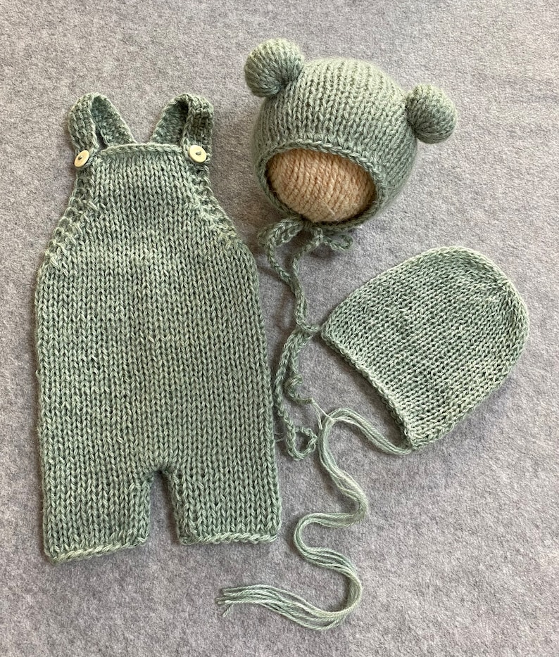 May include: A sage green knitted baby outfit set with a hat, a bonnet, and overalls. The hat has two small pom-poms on top.