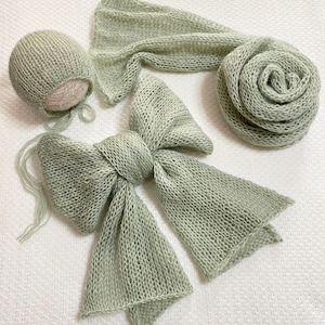 Puede incluir: Un conjunto de tres accesorios de fotografía para recién nacidos de punto verde salvia. El conjunto incluye un gorro, una envoltura y un lazo.