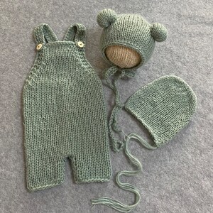 May include: A sage green knitted baby outfit set with a hat and a bonnet. The hat has bear ears and the bonnet has a tie.