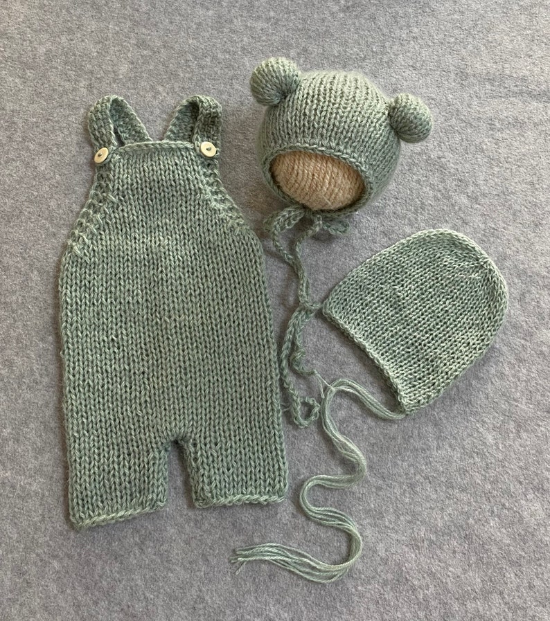 May include: A sage green knitted baby outfit set with a hat and a bonnet. The hat has bear ears and the bonnet has a tie.