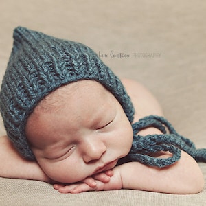 以下が含まれることがあります： 薄いベージュの布の上に寝ている新生児。ターコイズブルーのニット帽をかぶっています。帽子の紐は長く、床に垂れ下がっています。