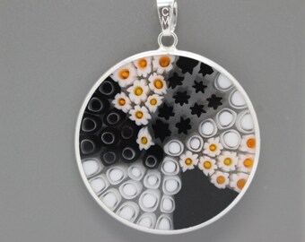May include: A round silver pendant with a black, white, and orange glass design. The design features a variety of shapes, including circles, stars, and flowers.