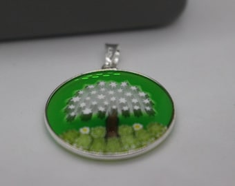 May include: A silver pendant with a green background and a white tree with white flowers. The tree is surrounded by green grass and white flowers.