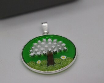 May include: A silver pendant with a green and white glass design of a tree with white flowers and a brown trunk. The pendant is round and has a silver bail.