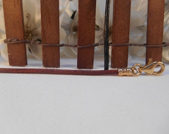 May include: A brown leather cord with a gold clasp. The cord is lying on a white surface in front of a wooden fence.
