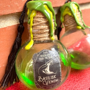 Puede incluir: Botella de vidrio verde con un tapón de corcho y un borla negra. La botella está llena de un líquido verde y tiene una etiqueta que dice "1809 Wardrath's Dark Unction Basilisk Venom 50 fl oz".