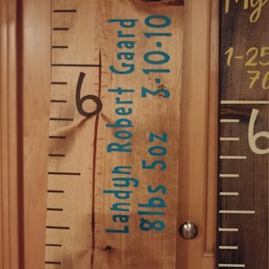 May include: Wooden growth chart ruler with black and teal lettering. The chart includes the name "Landyn Robert Gaard", birthdate, and weight. The ruler is marked with black lines and numbers.