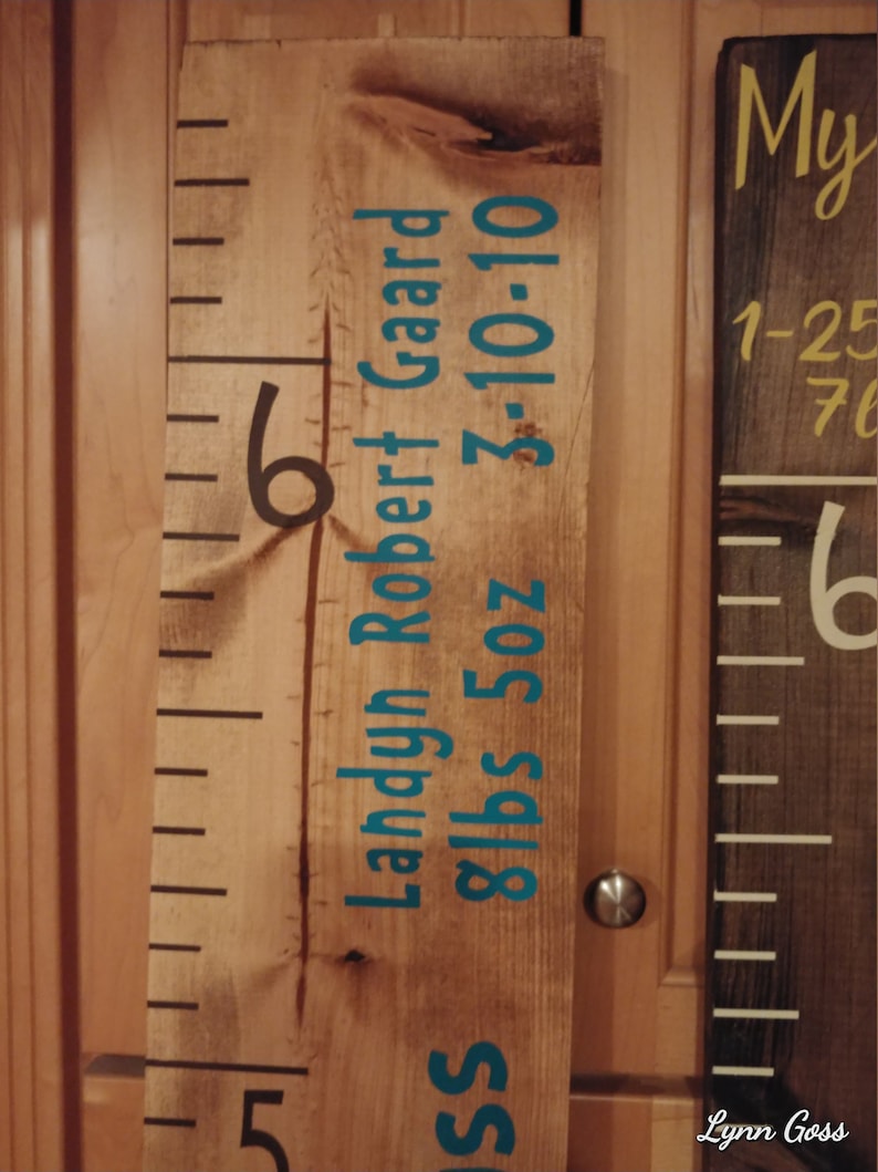 May include: Wooden growth chart ruler with black and teal lettering. The chart includes the name "Landyn Robert Gaard", birthdate, and weight. The ruler is marked with black lines and numbers.