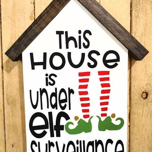 May include: A white wooden sign with a dark brown roof-shaped frame. The sign reads "This HOUSE is under ELF surveillance" in black lettering, with red and white striped legs and green elf shoes.