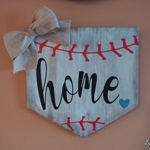 May include: A baseball-themed wooden sign with the word "home" in black script. The sign is painted light gray with red stitching details and a blue heart. A burlap bow is attached to the top.