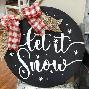 May include: A round, black wooden sign with the words "Let it Snow" in white script. The sign is decorated with white snowflakes and a red and white plaid bow with burlap accents. A festive winter decoration.