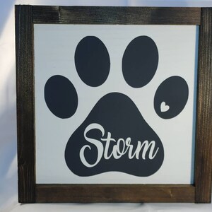 May include: A framed wooden sign with a white background features a large black paw print. The name "Storm" is written in cursive within the paw print. A small white heart is next to the paw print. The frame is dark brown.