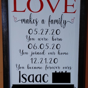 May include: Wooden framed sign with the words "LOVE makes a family" in red and black script. The sign includes dates and the name "Isaac". A decorative piece for a home.