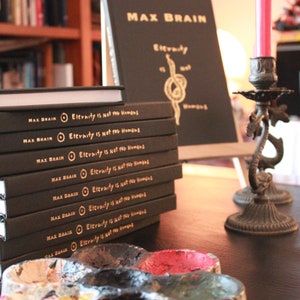 May include: A stack of black hardcover books with gold lettering on the spine. The title of the book is "Eternity is not for Humans". The books are on a wooden table with a black paint palette and a candle holder.