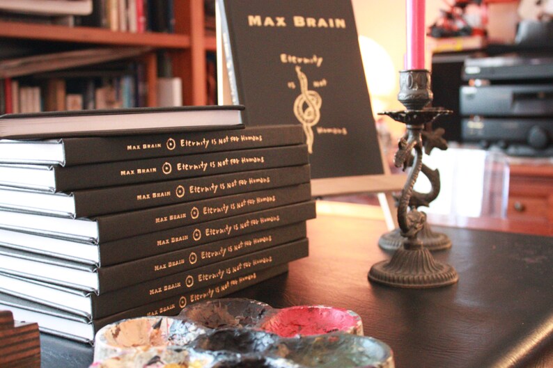 May include: A stack of black hardcover books with gold lettering on the spine. The title of the book is "Eternity is not for Humans". The books are on a wooden table with a black paint palette and a candle holder.