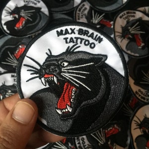 Op de afbeelding: Zwart-witte geborduurde patch met een knorrende panter en de tekst "Max Brain Tattoo".