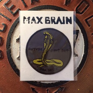 Può includere: Una toppa con la scritta "MAX BRAIN" in alto. La toppa presenta un cobra giallo su uno sfondo circolare grigio scuro. Il testo "FUTURE IS NOT SET" è sotto il cobra. La toppa è su una carta bianca.