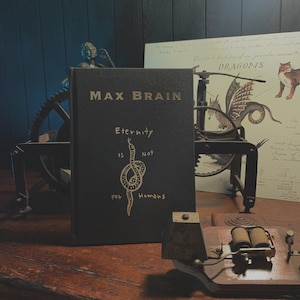 May include: A black hardcover book with the title "Max Brain" and the text "Eternity is Not for Humans" in gold lettering. The book is sitting on a wooden table with a vintage-looking machine and a small statue in the background.