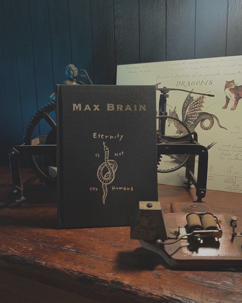May include: A black hardcover book with the title "Max Brain" and the text "Eternity is Not for Humans" in gold lettering. The book is sitting on a wooden table with a vintage-looking machine and a small statue in the background.