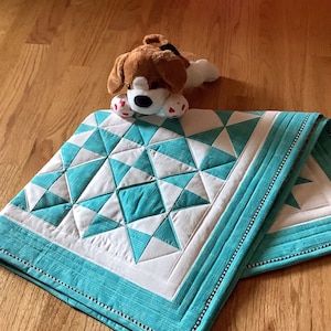 May include: A turquoise and white quilt with a geometric pattern. The quilt is folded in half and a small stuffed dog is lying on top of it.