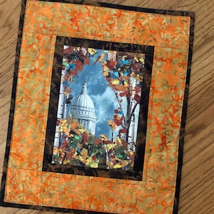 May include: A framed print of a building with a dome, surrounded by fall foliage. The print is set against a background of orange and brown fabric with a leaf pattern.