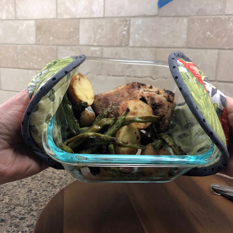 May include: A glass baking dish with a blue rim, filled with green beans, potatoes, and a piece of roasted meat. The dish is being held by two green and gray oven mitts.